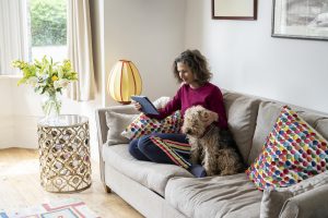 Mature caucasian female sits on couch with her pet Welsh Terrier looking at social media