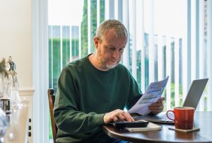 Portrait of a mid adult man in his 30s checking his bills at home. He has a worried expression and touches his face with his hand while looking at the bills. He is surrounded by documents, his laptop and a calculator.